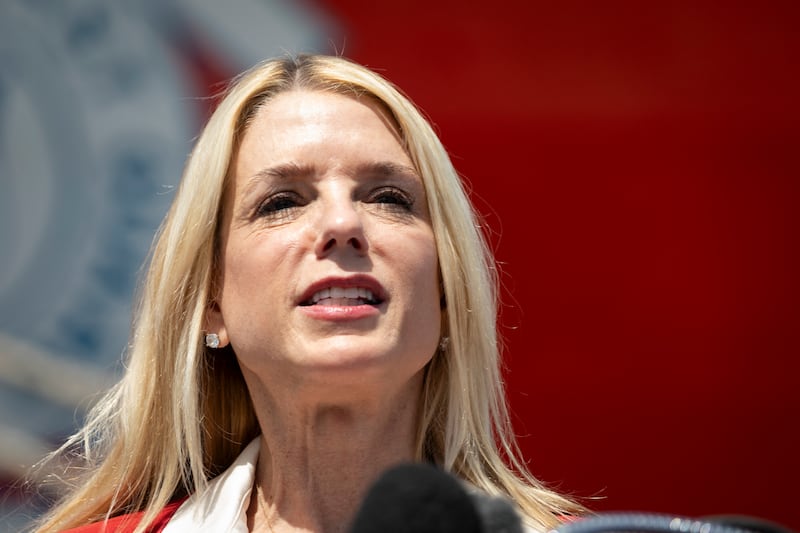 US Attorney General Pam Bondi stands by a podium and squints in the sunlight.