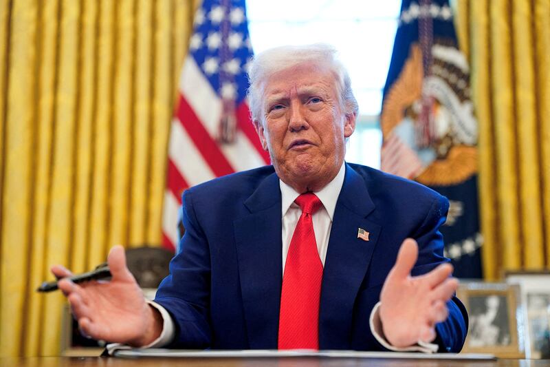 Donald Trump gestures with open hands and what appears to be a sneering expression. He is photographed from the chest up, clearly behind the Resolute Desk in the Oval Office, with U.S flag on his right and the official seal on a flag on his left.