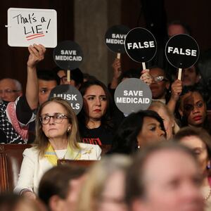 Democrats hold protest signs as Trump speaks.