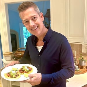 Sean Duffy in a kitchen in front of a white countertop and white wooden cabinets holding a plate with two tacos. He is wearing a dark blue untucked shirt with a low-cut t underneath and lighter casual trousers.