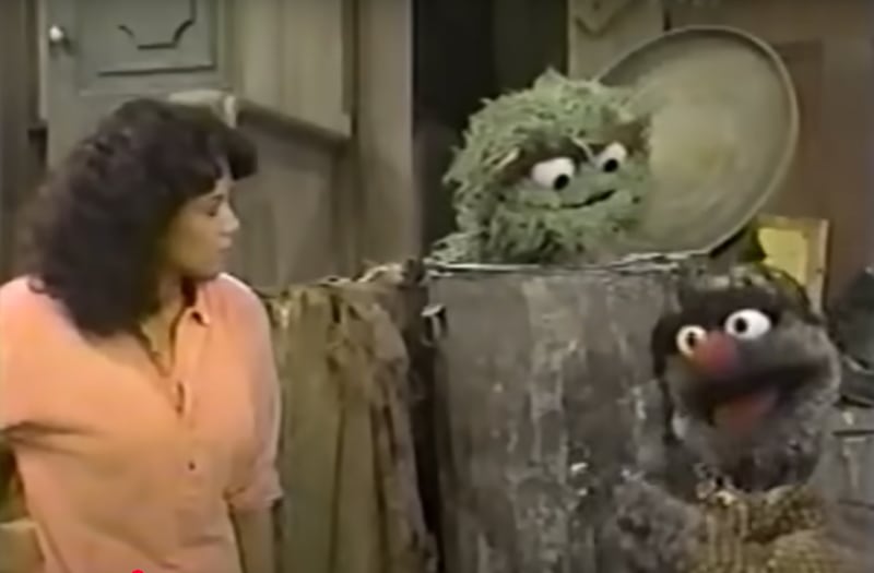 On Sesame Street, a green puppet in a trash can and a person look at a grey puppet that is speaking.