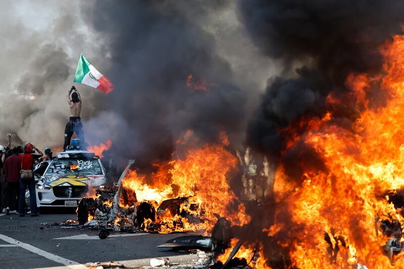 LOS ANGELES, LOS ANGELES - JUNE 08: A protestor holds up a Mexican flag as burning cars line the street on June 08, 2025 in Los Angeles, California. Tensions in the city remain high after the Trump administration called in the National Guard against the wishes of city leaders following two days of clashes with police during a series of immigration raids.  (Photo by Mario Tama/Getty Images)