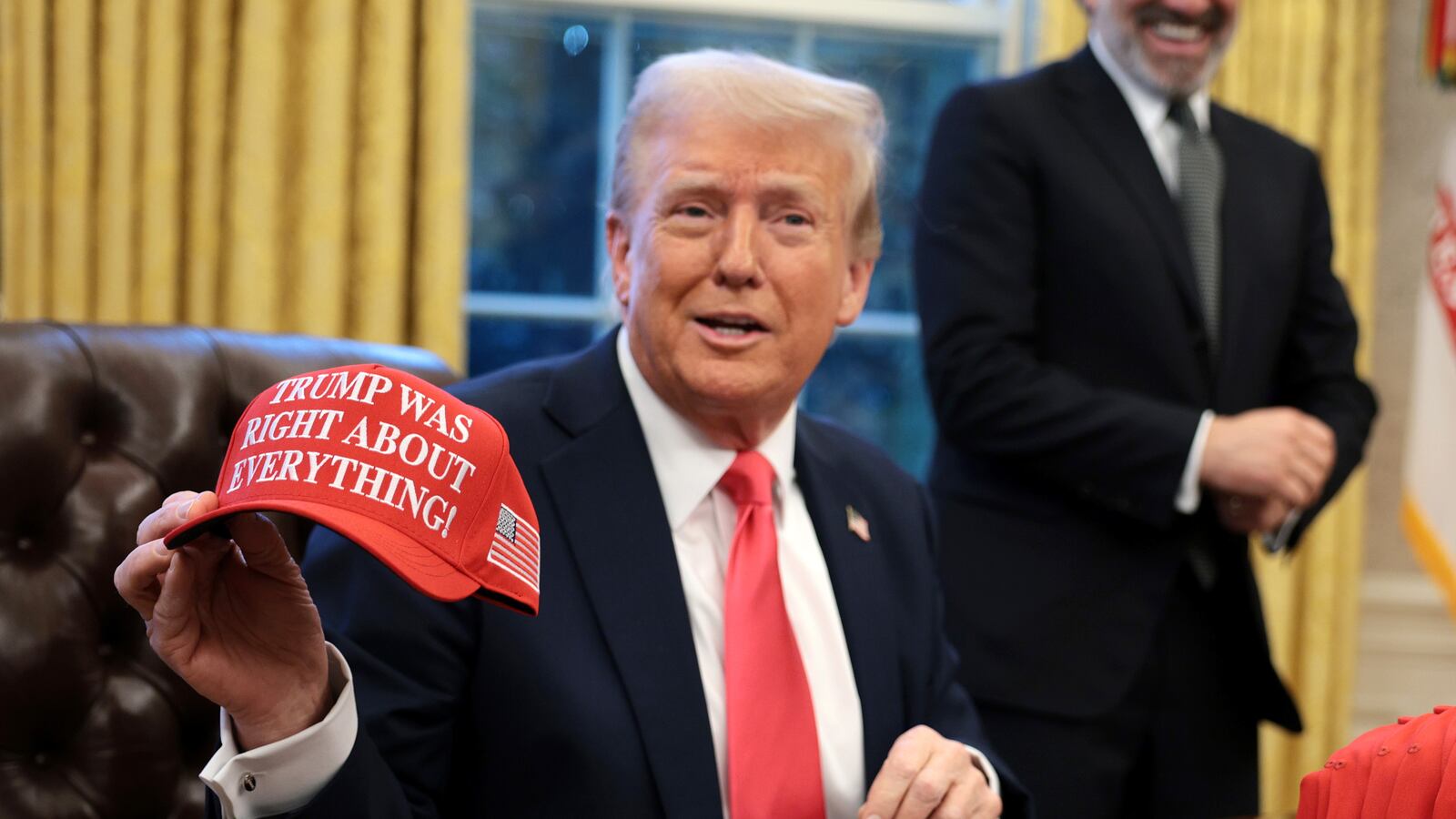 President Donald Trump sits at his desk in the Oval Office and holds up a hat that says "Trump was right about everything."