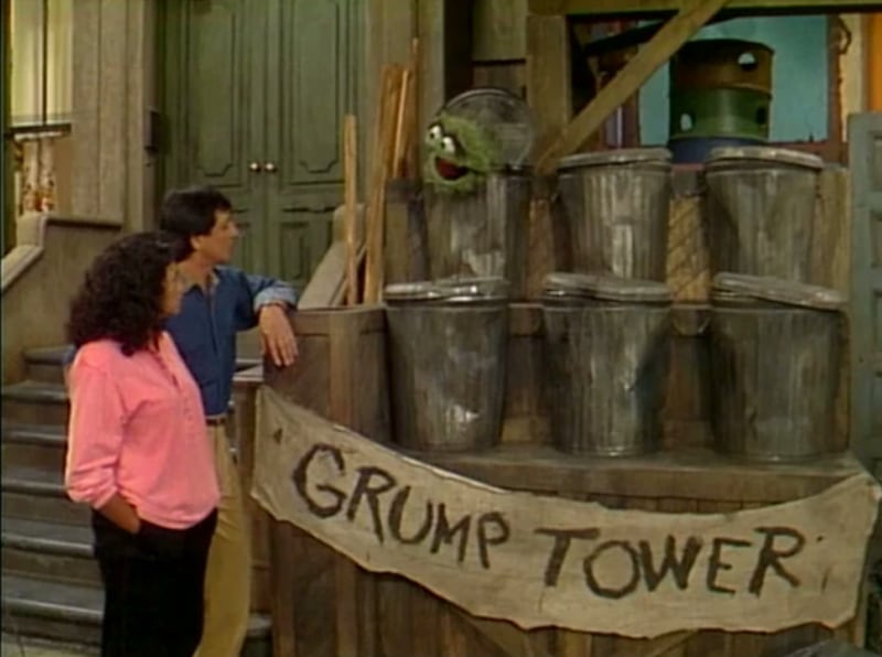 On Sesame Street, two people look at a green Muppet whose head is coming out of a trash can. There are two rows of three trash cans beside the stoop. In front is a "Grump Tower" sign