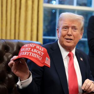 President Donald Trump sits at his desk in the Oval Office and holds up a hat that says "Trump was right about everything."