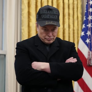 Elon Musk looks down with his arms folded while wearing a MAGA hat in the Oval Office.